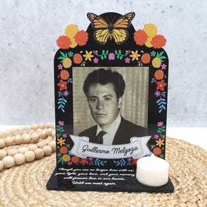 May include: A black memorial plaque featuring a monarch butterfly, colorful marigold flowers, and a black-and-white portrait of Guillermo Melgoza. The base includes a white tea light candle and text that reads, Though you are no longer here with us.
