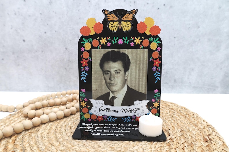 May include: A black memorial plaque featuring a monarch butterfly, colorful marigold flowers, and a black-and-white portrait of Guillermo Melgoza. The base includes a white tea light candle and text that reads, Though you are no longer here with us.