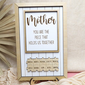 Puede incluir: Un marco de madera con fondo blanco y el texto "Madre T&uacute; eres la pieza que nos mantiene unidos". Debajo del texto hay piezas de rompecabezas de madera con nombres en ellas: Andrew, Andres, Mother, Ashley, Miguel, Kryla, Brenda, Dom, Jacob, Junior.