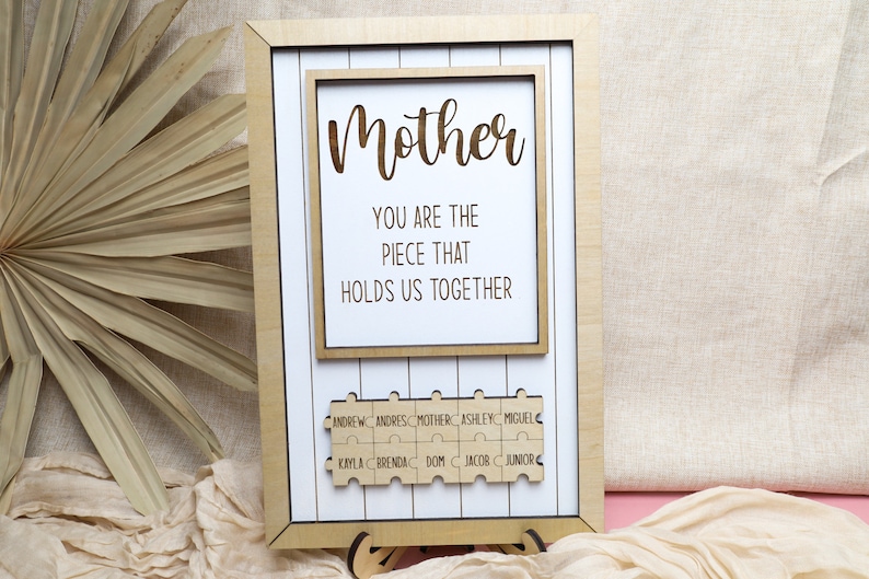 Puede incluir: Un marco de madera con fondo blanco y el texto "Madre T&uacute; eres la pieza que nos mantiene unidos". Debajo del texto hay piezas de rompecabezas de madera con nombres en ellas: Andrew, Andres, Mother, Ashley, Miguel, Kryla, Brenda, Dom, Jacob, Junior.