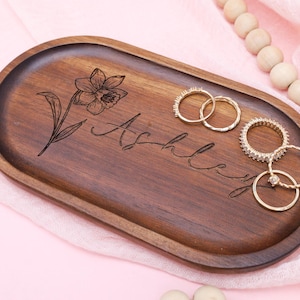 May include: A wooden, oval-shaped tray with a floral engraving and the name "Ashley" in cursive. Several gold rings are placed on the tray. The tray is on a pink surface with a beaded garland.
