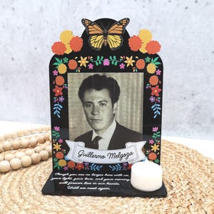 May include: A black acrylic memorial plaque featuring a monarch butterfly, colorful marigold flowers, and a black and white portrait of a man named Guillermo Melgoza. The base includes a white tea light candle and a memorial inscription in white text.