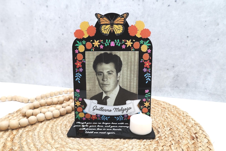 May include: A black acrylic memorial plaque featuring a monarch butterfly, colorful marigold flowers, and a black and white portrait of a man named Guillermo Melgoza. The base includes a white tea light candle and a memorial inscription in white text.