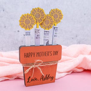 Op de afbeelding: Houten plantenbak met de tekst Happy Mother's Day en de woorden Love, Ashley. De plantenbak is versierd met vier gele papieren zonnebloemen en bevat geld.