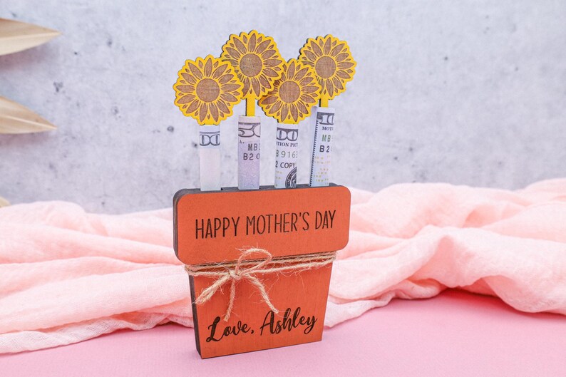 Op de afbeelding: Houten plantenbak met de tekst Happy Mother's Day en de woorden Love, Ashley. De plantenbak is versierd met vier gele papieren zonnebloemen en bevat geld.