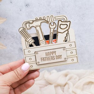 May include: A wooden toolbox-shaped card holder with cut-out tools, including a hammer, screwdriver, saw, and wrench. The words "HAPPY FATHERS DAY" are engraved on a rectangular plaque on the front of the toolbox.