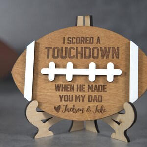 May include: Wooden football-shaped sign with the text "I scored a touchdown when he made you my dad" and a heart with the names "Jackson & Jake".