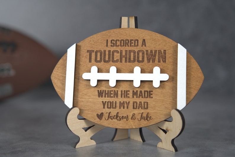 May include: Wooden football-shaped sign with the text "I scored a touchdown when he made you my dad" and a heart with the names "Jackson & Jake".