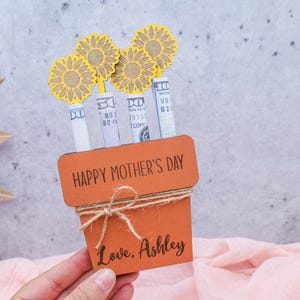 Op de afbeelding: Een bruine houten plantenbak met de tekst "Happy Mother's Day" en "Love, Ashley" erop. De plantenbak is versierd met gele papieren bloemen en briefjes.