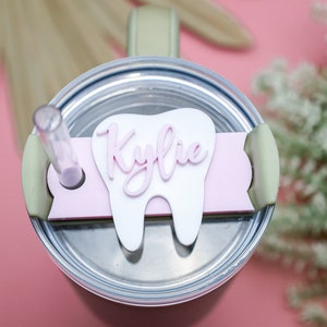 May include: A white tooth-shaped lid with a pink name "Kylie" on it. The lid is on top of a stainless steel tumbler with a green lid and a clear straw.