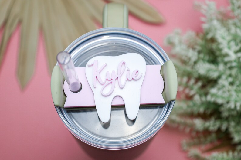 May include: A white tooth-shaped lid with a pink name "Kylie" on it. The lid is on top of a stainless steel tumbler with a green lid and a clear straw.
