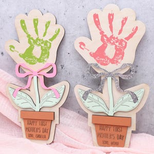 May include: Two wooden flower crafts with handprints in the center. The flowers are green and pink with a pink and silver bow. The pots are brown with the text "Happy First Mother's Day" and the child's name.