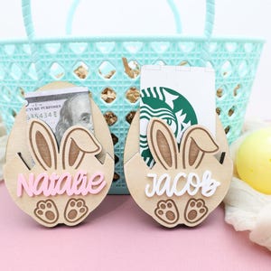 May include: Wooden Easter egg holders with bunny ears, personalized with the names "Natalie" and "Jacob" in pink and white. One holds a $100 bill, the other a gift card. A light blue basket is in the background.