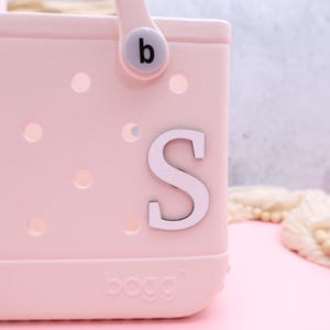 Pode incluir: Uma bolsa tote rosa com uma letra branca "S" presa na frente. A bolsa tem uma al&ccedil;a branca com uma letra preta "b" nela. A bolsa tem uma etiqueta branca que diz "BOGO" na parte inferior.