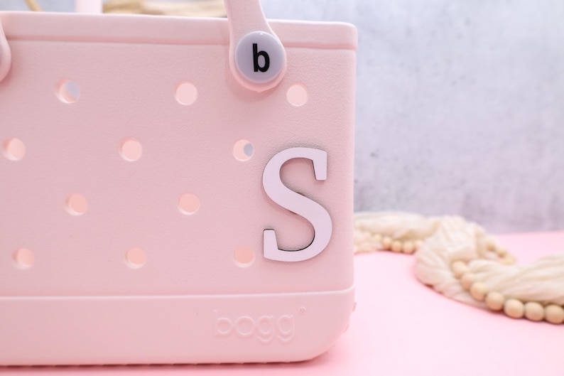 Pode incluir: Uma bolsa tote rosa com uma letra branca "S" presa na frente. A bolsa tem uma al&ccedil;a branca com uma letra preta "b" nela. A bolsa tem uma etiqueta branca que diz "BOGO" na parte inferior.