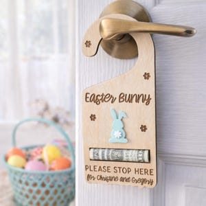 Puede incluir: Colgador de puerta de madera de conejito de Pascua con un conejito azul claro y diseño de flores. El texto "Easter Bunny" y "PLEASE STOP HERE for Christine and Gregory" están grabados en la madera. Una ranura contiene un billete enrollado.