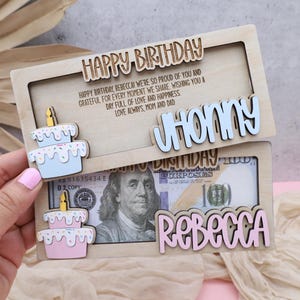 Puede incluir: Una tarjeta de cumplea&ntilde;os de madera con una ranura para dinero en efectivo. La tarjeta tiene un pastel azul y blanco con chispas y el nombre "JHONNY" en la mitad superior. La mitad inferior tiene un pastel rosa y blanco con chispas y el nombre "REBECCA" y un billete de d&oacute;lar con el retrato de un hombre.