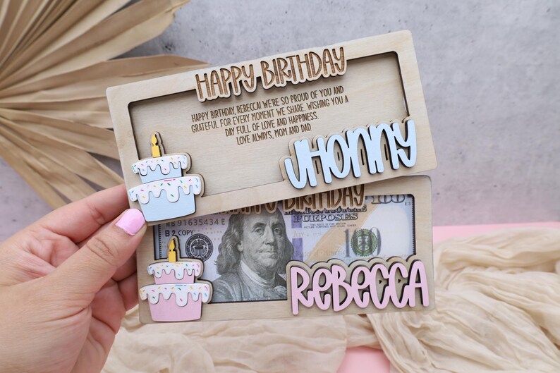 Puede incluir: Una tarjeta de cumplea&ntilde;os de madera con una ranura para dinero en efectivo. La tarjeta tiene un pastel azul y blanco con chispas y el nombre "JHONNY" en la mitad superior. La mitad inferior tiene un pastel rosa y blanco con chispas y el nombre "REBECCA" y un billete de d&oacute;lar con el retrato de un hombre.