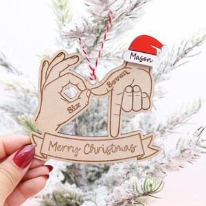 May include: Wooden Christmas ornament with a hand gesture design. The ornament features two hands forming the numbers six and seven, a Santa hat with the name "Mason", and a banner that reads "Merry Christmas".