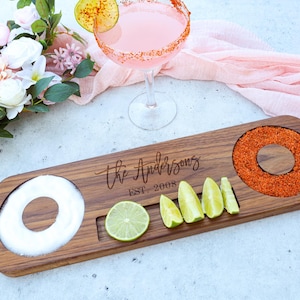 May include: A wooden cocktail serving board with a salt rim, chili rim, and lime wedges. The board is engraved with "The Andersons EST. 2008". A pink cocktail with a lime garnish sits nearby, along with a bouquet of flowers.