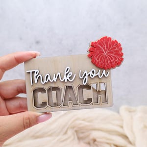 May include: A wooden card with the words "Thank you COACH" in white and a red pom-pom accent. The card is made of light-colored wood and has a cutout design for the word "COACH".