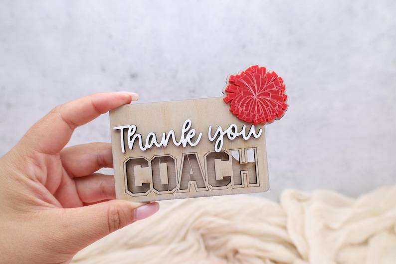 May include: A wooden card with the words "Thank you COACH" in white and a red pom-pom accent. The card is made of light-colored wood and has a cutout design for the word "COACH".