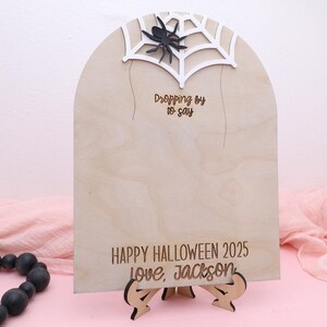 May include: A wooden Halloween sign with a spider web and spider design. The sign reads "Dropping by to say" and "Happy Halloween 2025 love, Jackson". The sign is supported by a wooden stand.