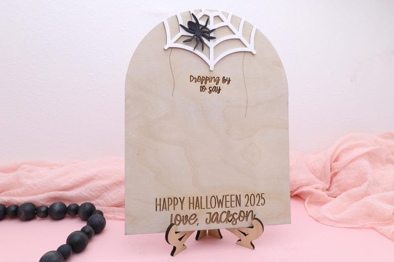 May include: A wooden Halloween sign with a spider web and spider design. The sign reads "Dropping by to say" and "Happy Halloween 2025 love, Jackson". The sign is supported by a wooden stand.