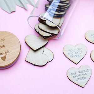 May include: A glass jar filled with wooden heart-shaped tokens with handwritten messages on them. The jar has a wooden lid with the text "I Love You Because..." on it. The jar is on a pink surface with several of the wooden hearts scattered around it.
