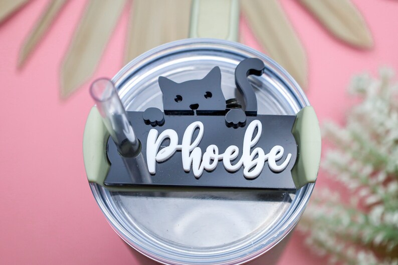May include: Black and white cat silhouette with a white name tag that reads "Phoebe" on a black background. The name tag is attached to a green lid of a stainless steel tumbler.