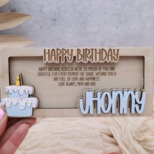 Puede incluir: Una tarjeta de cumplea&ntilde;os de madera con un recorte de un pastel y el texto "HAPPY BIRTHDAY". La tarjeta tambi&eacute;n incluye el texto "HAPPY BIRTHDAY, REBECCA! WE'RE SO PROUD OF YOU AND GRATEFUL FOR EVERY MOMENT WE SHARE. WISHING YOU A DAY FULL OF LOVE AND HAPPINESS. LOVE ALWAYS, MOM AND DAD." y el nombre "JHONNY" en una fuente recortada.