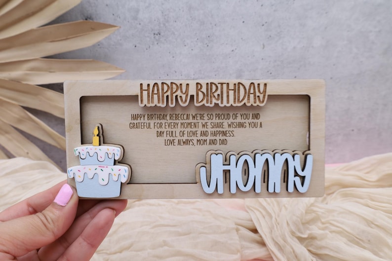 Puede incluir: Una tarjeta de cumplea&ntilde;os de madera con un recorte de un pastel y el texto "HAPPY BIRTHDAY". La tarjeta tambi&eacute;n incluye el texto "HAPPY BIRTHDAY, REBECCA! WE'RE SO PROUD OF YOU AND GRATEFUL FOR EVERY MOMENT WE SHARE. WISHING YOU A DAY FULL OF LOVE AND HAPPINESS. LOVE ALWAYS, MOM AND DAD." y el nombre "JHONNY" en una fuente recortada.