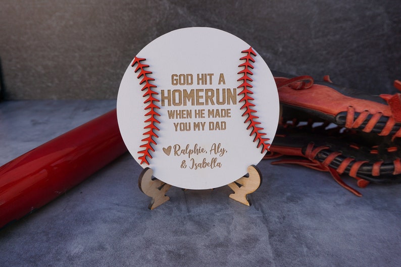 May include: White wooden baseball-shaped sign with red stitching. The sign reads "God hit a homerun when he made you my dad."  The sign is on a small wooden stand. The names "Ralphie, Aly, & Isabella" are written below the text.