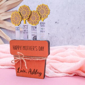 Op de afbeelding: Een houten plantenbak met de woorden "Happy Mother's Day" en "Love, Ashley" erop gegraveerd. De plantenbak is versierd met vier gele papieren bloemen en bankbiljetten die in de stelen zijn gestoken.
