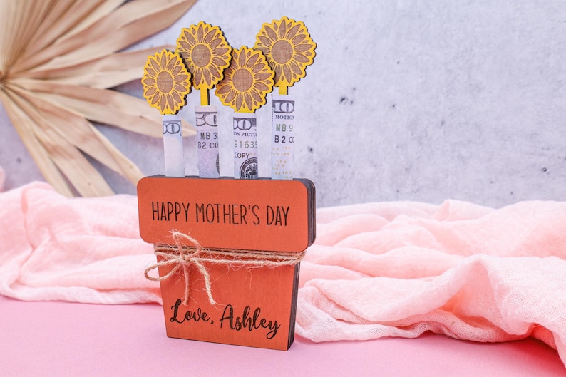 Op de afbeelding: Een houten plantenbak met de woorden "Happy Mother's Day" en "Love, Ashley" erop gegraveerd. De plantenbak is versierd met vier gele papieren bloemen en bankbiljetten die in de stelen zijn gestoken.