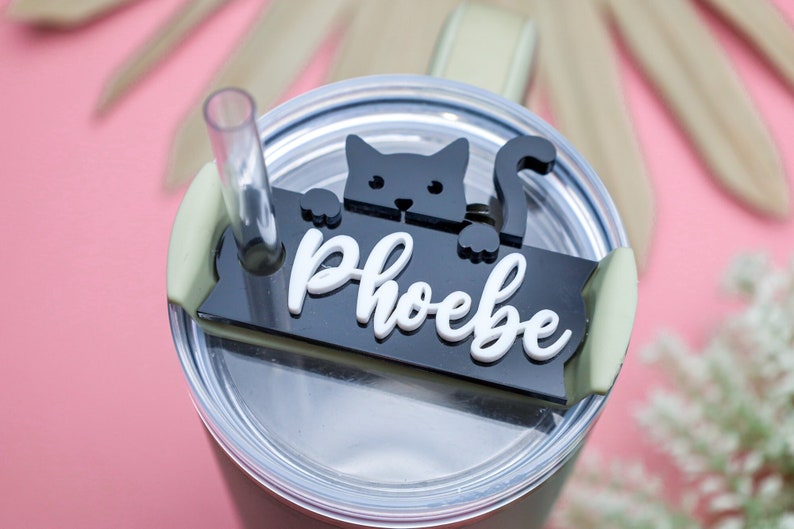 May include: Black and white cat silhouette with a white name tag that reads "Phoebe" on a black background. The name tag is attached to a clear lid for a tumbler.