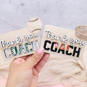 May include: Two wooden gift card holders with the words "Thank you COACH" in white script. One has a baseball graphic, the other a hockey puck. The word COACH is in green and black, and black and orange.