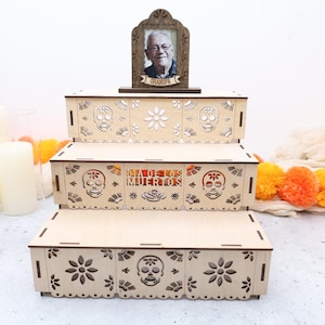 May include: A three-tiered wooden display stand featuring laser-cut sugar skull and flower motifs. The middle tier displays the text "DIA DE LOS MUERTOS" and the top tier holds a framed photograph of an elderly man with the label "GRANDPA".