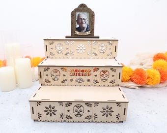 May include: A three-tiered wooden display stand featuring laser-cut sugar skull and flower motifs. The middle tier displays the text "DIA DE LOS MUERTOS" and the top tier holds a framed photograph of an elderly man with the label "GRANDPA".