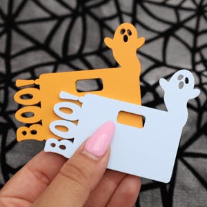 May include: Two Halloween-themed ghost-shaped ornaments. One is orange with the word "BOO!" written on it. The other is light blue with a ghost shape on it.