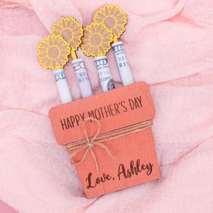 Op de afbeelding: Een houten plantenbak met de tekst "Happy Mother's Day" en "Love, Ashley" erop. De plantenbak is gevuld met opgerolde dollarbiljetten en versierd met gele papieren bloemen.
