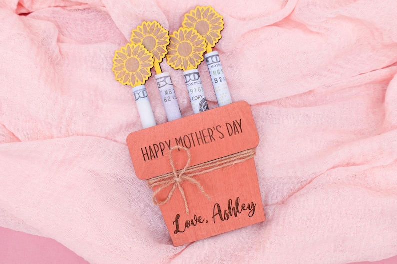 Op de afbeelding: Een houten plantenbak met de tekst "Happy Mother's Day" en "Love, Ashley" erop. De plantenbak is gevuld met opgerolde dollarbiljetten en versierd met gele papieren bloemen.