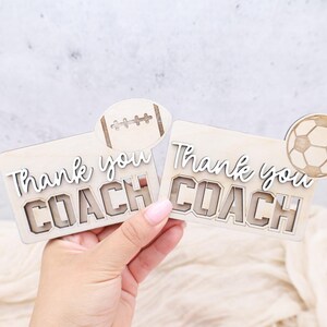 May include: Two wooden plaques with the words "Thank you COACH" in a layered design. One plaque has a football accent, the other a soccer ball. The plaques are light brown with white lettering.