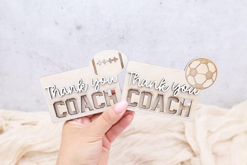 May include: Two wooden plaques with the words "Thank you COACH" in a layered design. One plaque has a football accent, the other a soccer ball. The plaques are light brown with white lettering.