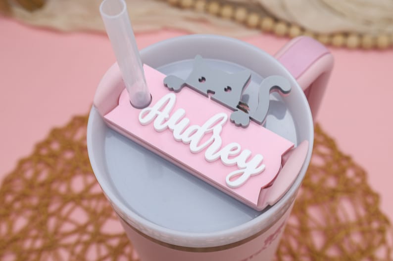 May include: Pink and gray personalized tumbler with a cat design and the name "Audrey" on a pink name tag.