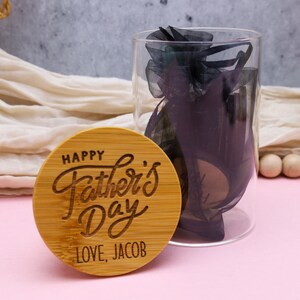 May include: A round wooden plaque with the text "Happy Father's Day" in a cursive font. The plaque also has the text "Love, Jacob" engraved on it. The plaque is sitting on a pink surface.