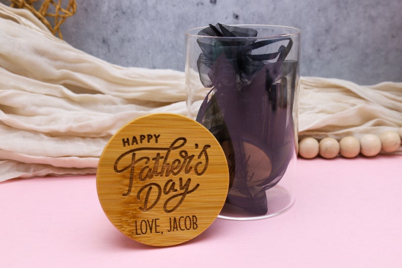 May include: A round wooden plaque with the text "Happy Father's Day" in a cursive font. The plaque also has the text "Love, Jacob" engraved on it. The plaque is sitting on a pink surface.