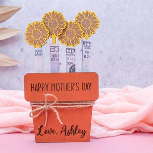 Op de afbeelding: Een houten plantenbak met de woorden "Happy Mother's Day" en "Love, Ashley" erop gegraveerd. De plantenbak is versierd met vier gele papieren zonnebloemen en heeft een bruine touw strik aan de bovenkant.