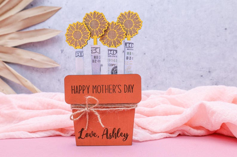 Op de afbeelding: Een houten plantenbak met de woorden "Happy Mother's Day" en "Love, Ashley" erop gegraveerd. De plantenbak is versierd met vier gele papieren zonnebloemen en heeft een bruine touw strik aan de bovenkant.