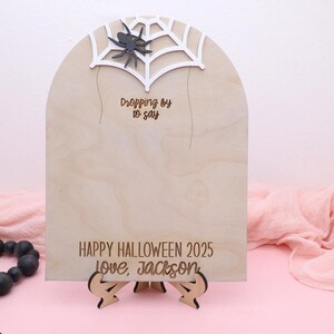 May include: A wooden Halloween sign with a spider web and spider design. The sign reads "Dropping by to say" and "Happy Halloween 2025 Love, Jackson". The sign is supported by a wooden stand.